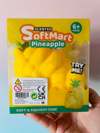A hand holding a boxed Scented SoftMart Pineapple toy against a pastel pink and blue background. The package is yellow with green and orange branding, shows a yellow pineapple-shaped squishy toy inside, and includes a 'TRY ME!' callout and age 6+ graphic.