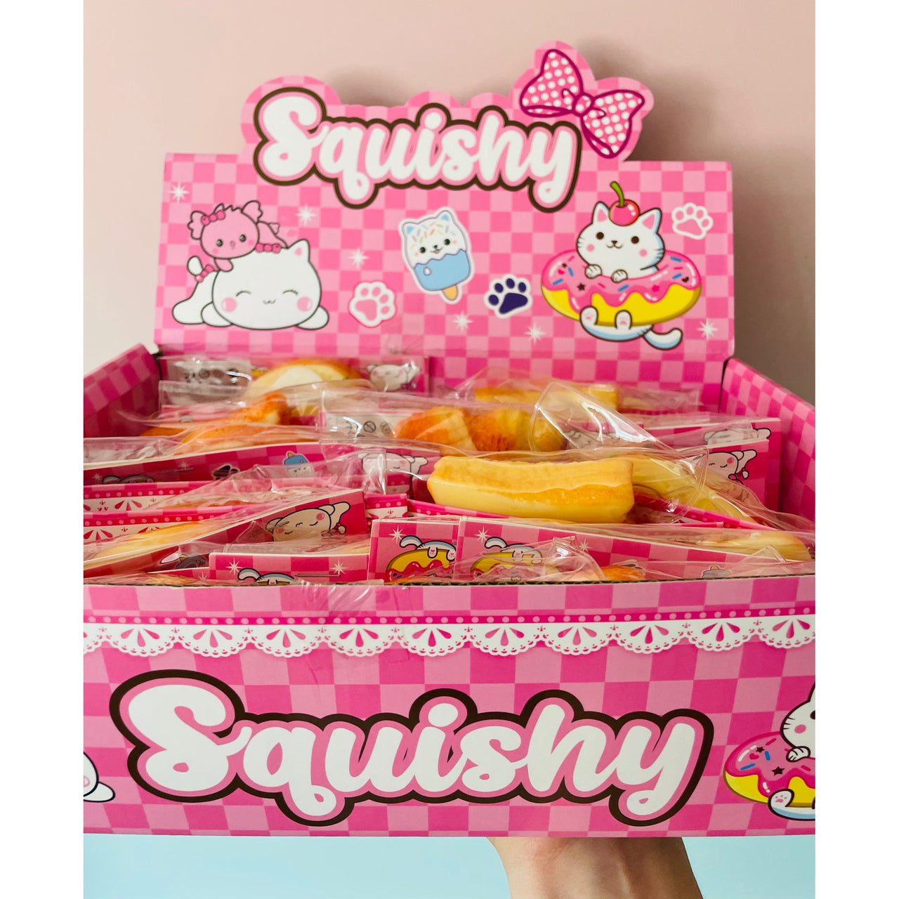A pink display box filled with individually wrapped squishy toys, with a large 'Squishy' sign on top. A hand is holding the box against a soft pink background.