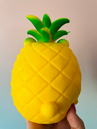 A bright yellow pineapple-shaped squishy toy being held in a hand against a soft beige and light blue gradient background.