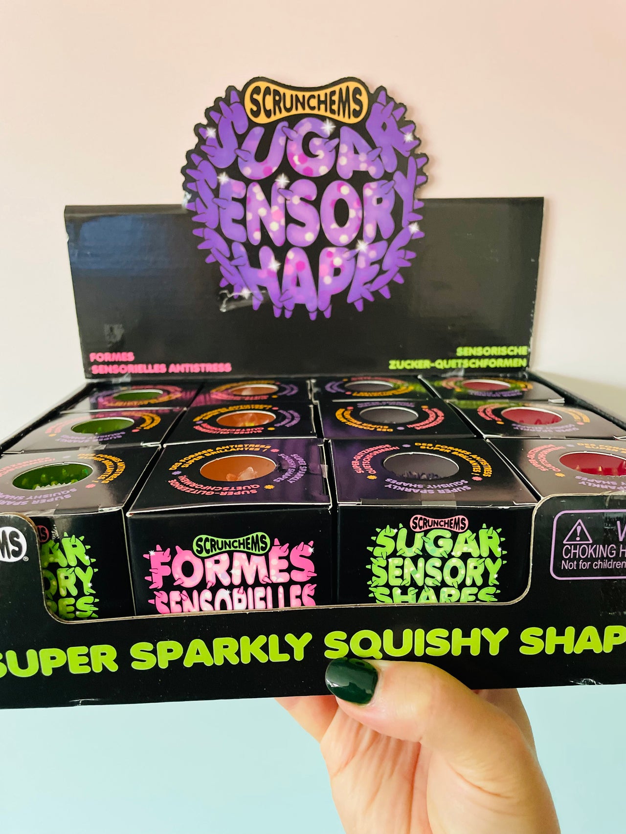 A hand with dark green nail polish holds a display box of Scrunchems Sugar Sensory Shapes. The packaging is black with vibrant purple, green, and pink text and graphics, showing multiple small cylindrical containers inside.