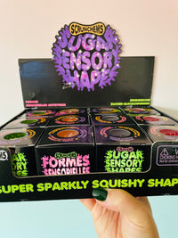 A hand with dark green nail polish holds a display box of Scrunchems Sugar Sensory Shapes. The packaging is black with vibrant purple, green, and pink text and graphics, showing multiple small cylindrical containers inside.