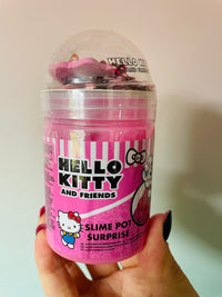 A hand holding a Hello Kitty and Friends Slime Pot Surprise toy container. The product is a pink plastic jar with a clear dome lid, containing pink and purple slime, with the Hello Kitty branding prominently displayed on the front label.