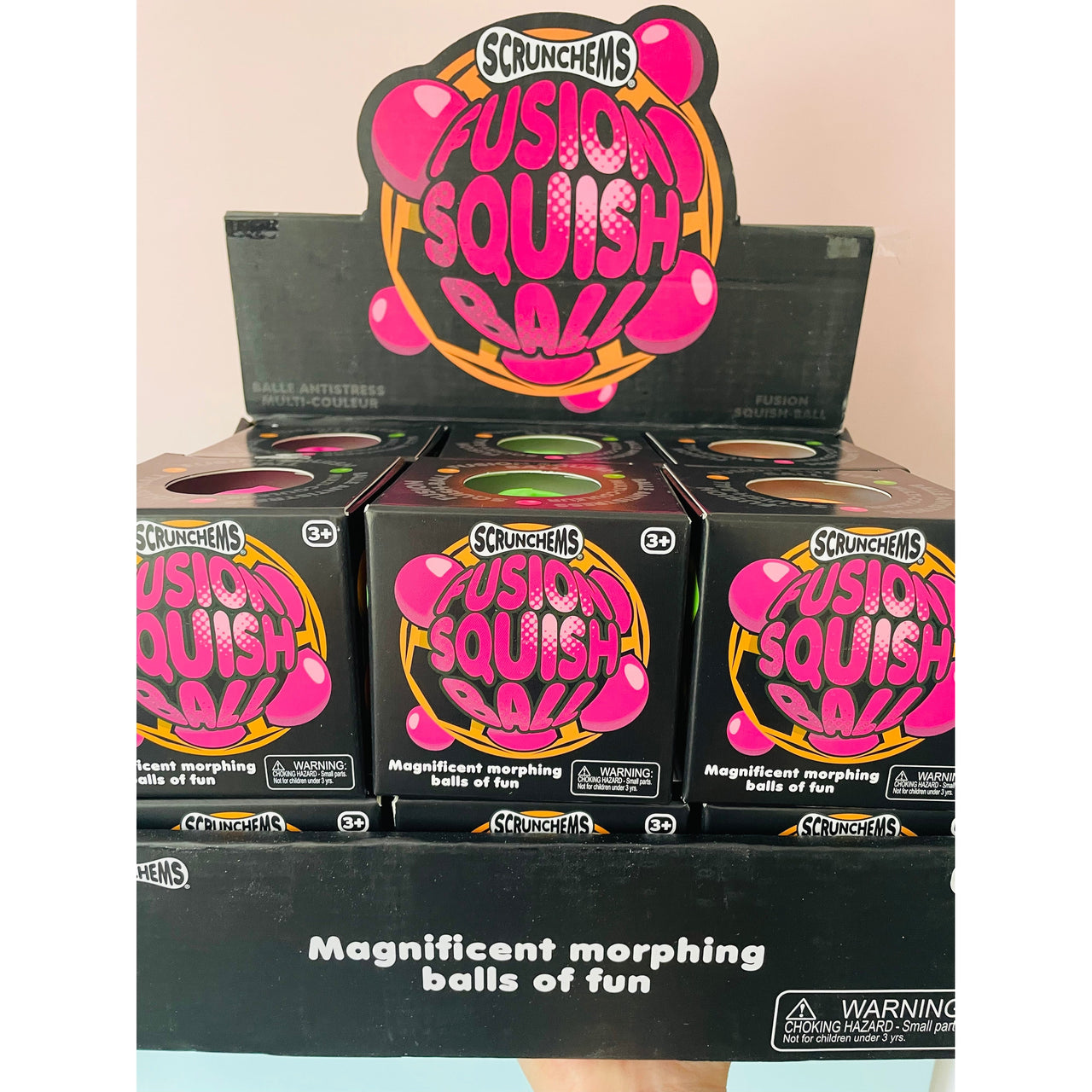 Scrunchems Fusion Squish Ball toy packaging display showing multiple black boxes with bright pink and yellow graphics, plus a larger sign above reading 'Scrunchems Fusion Squish Ball'.
