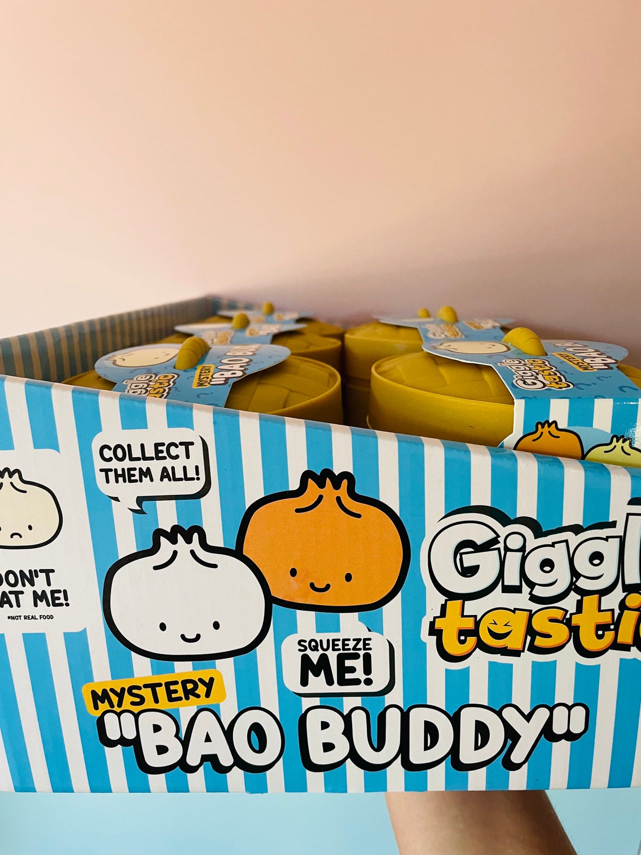 A box of Giggling Tasty 'Bao Buddy' mystery snack containers held by a hand against a soft pink background. The packaging features blue and white stripes with cartoon dumpling graphics and text like 'Collect them all!' and 'Squeeze me!'.