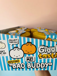 A box of Giggling Tasty 'Bao Buddy' mystery snack containers held by a hand against a soft pink background. The packaging features blue and white stripes with cartoon dumpling graphics and text like 'Collect them all!' and 'Squeeze me!'.