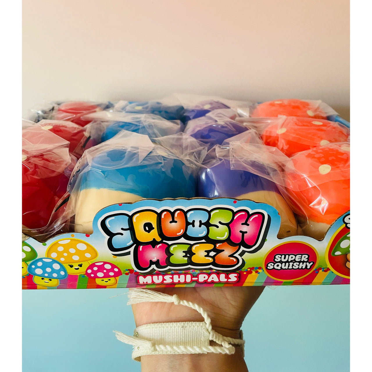 A hand holding a box of colorful Squish Meez Mushi-Pals squishy toys, showing multiple pastel-colored mushroom-shaped squishies wrapped in clear plastic inside a branded box.