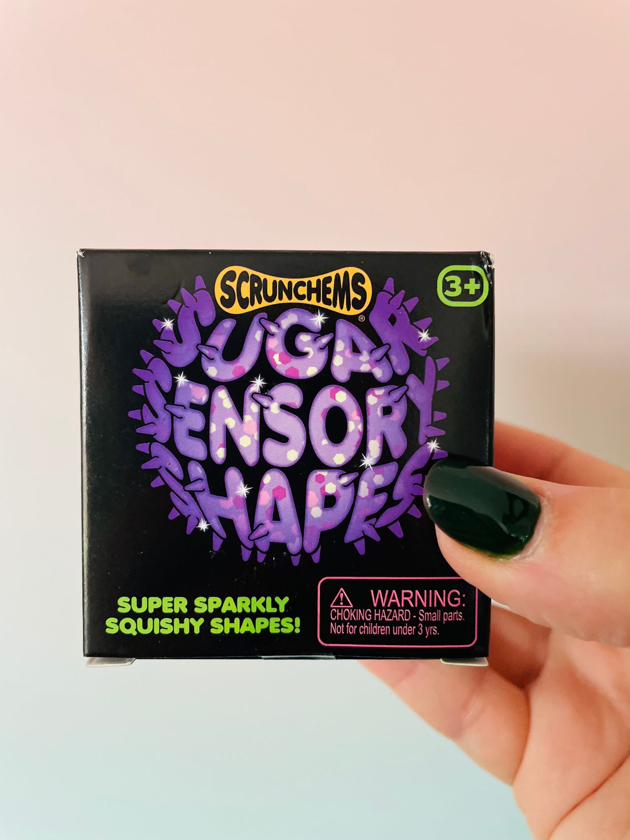 A hand with dark green nail polish holds a black box of Scrunchems Sugar Sensor Shapes squishy toys against a soft pink and light blue gradient background.