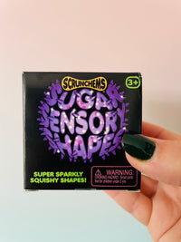 A hand with dark green nail polish holds a black box of Scrunchems Sugar Sensor Shapes squishy toys against a soft pink and light blue gradient background.