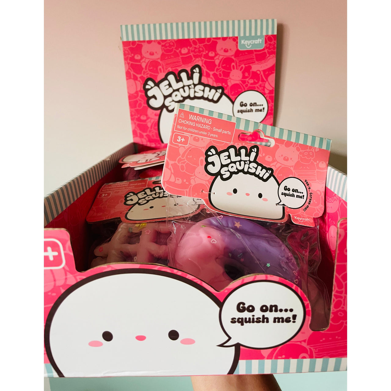 A display box filled with Jellii Squishies, including a pink and purple donut-shaped squishy toy, packaged in pink and white packaging with cartoon graphics.