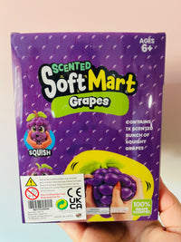 A hand holding a purple package of Scented SoftMart Grapes. The packaging features a purple grape pattern, a cartoon grape character, and a yellow graphic of a hand squeezing a grape.