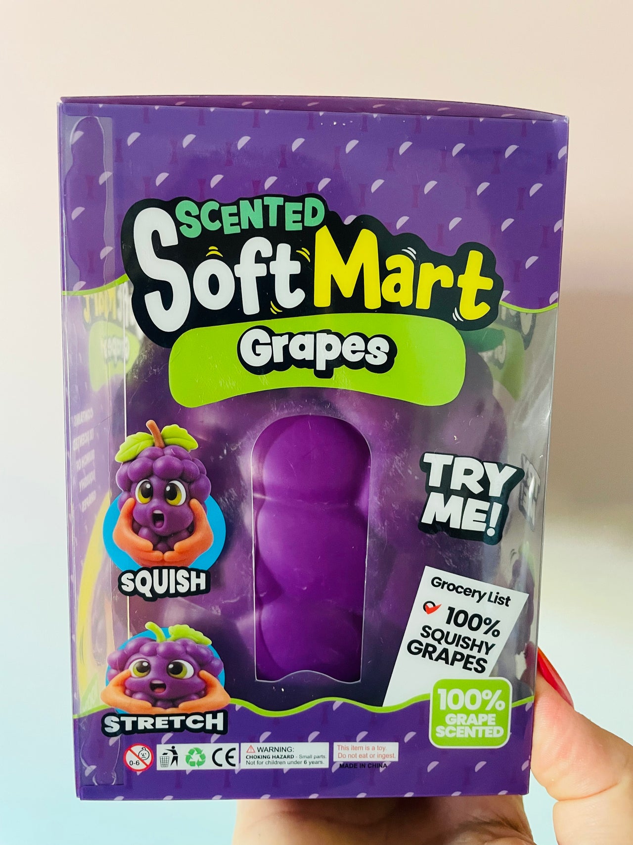 A hand holding a purple box of SoftMart Scented Soft Grapes. The packaging features a clear window showing a purple, squishy grape-shaped toy inside, with cartoon illustrations of a squishy grape and a stretchy grape.
