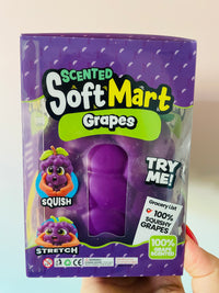 A hand holding a purple box of SoftMart Scented Soft Grapes. The packaging features a clear window showing a purple, squishy grape-shaped toy inside, with cartoon illustrations of a squishy grape and a stretchy grape.