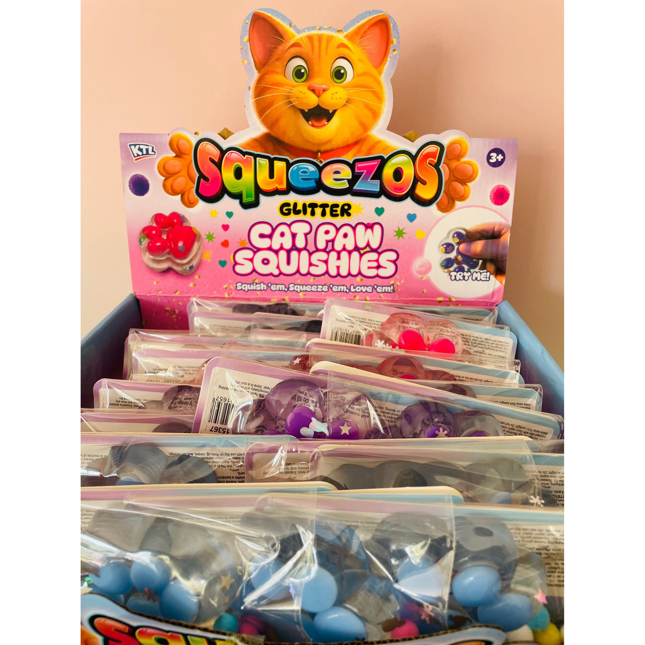 A display box filled with packaged Squeezos Glitter Cat Paw Squishies, showing multiple clear bags of blue, purple, and pink squishies in front of a colorful retail box with a cartoon cat face.