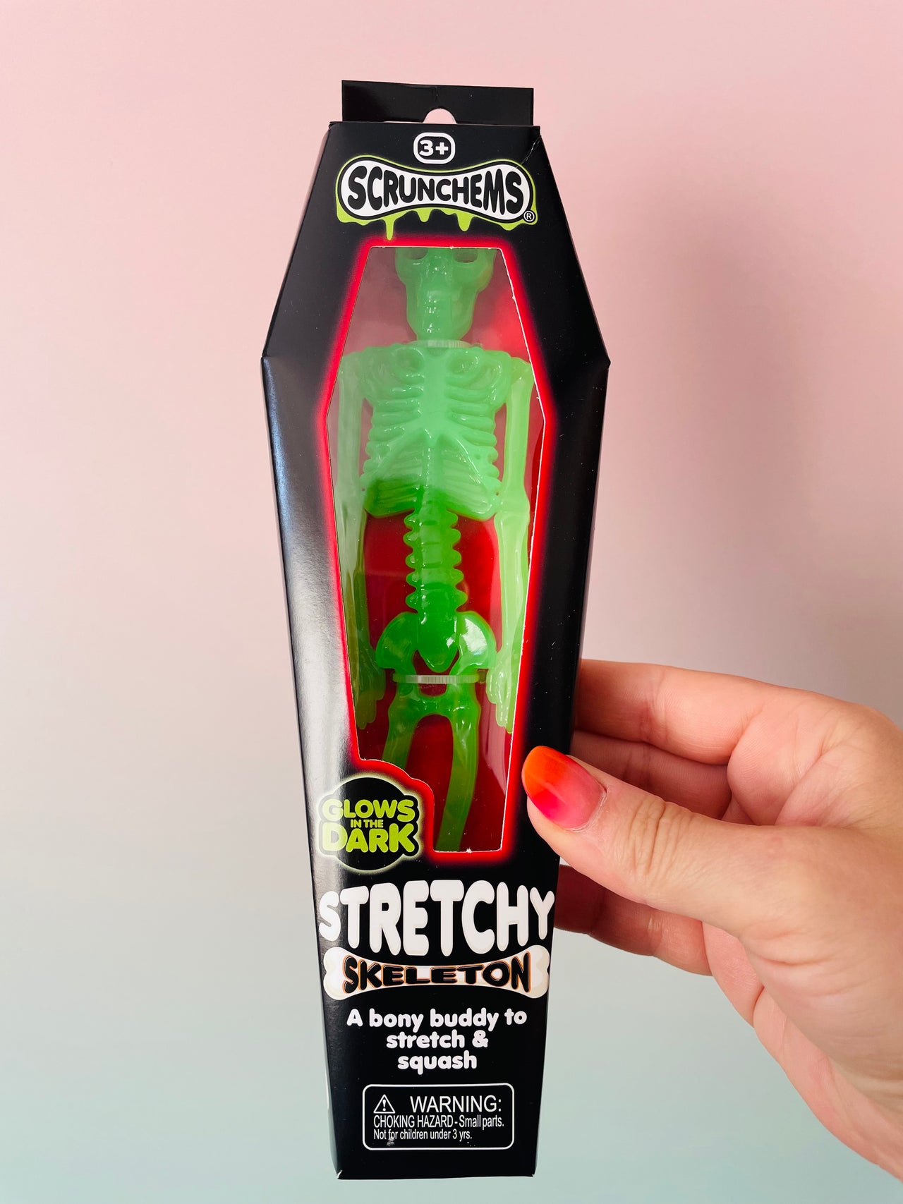 A hand holds a packaged Scrunchems Stretchy Skeleton toy. The product is in its black coffin-shaped retail box with a clear window showing a bright green glow-in-the-dark skeleton figure inside.