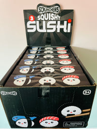 A display box of Scrunchems 3 Squishy Sushi toys, showing multiple individual packaged sushi-shaped squishies inside a black cardboard display box with cartoon sushi illustrations.