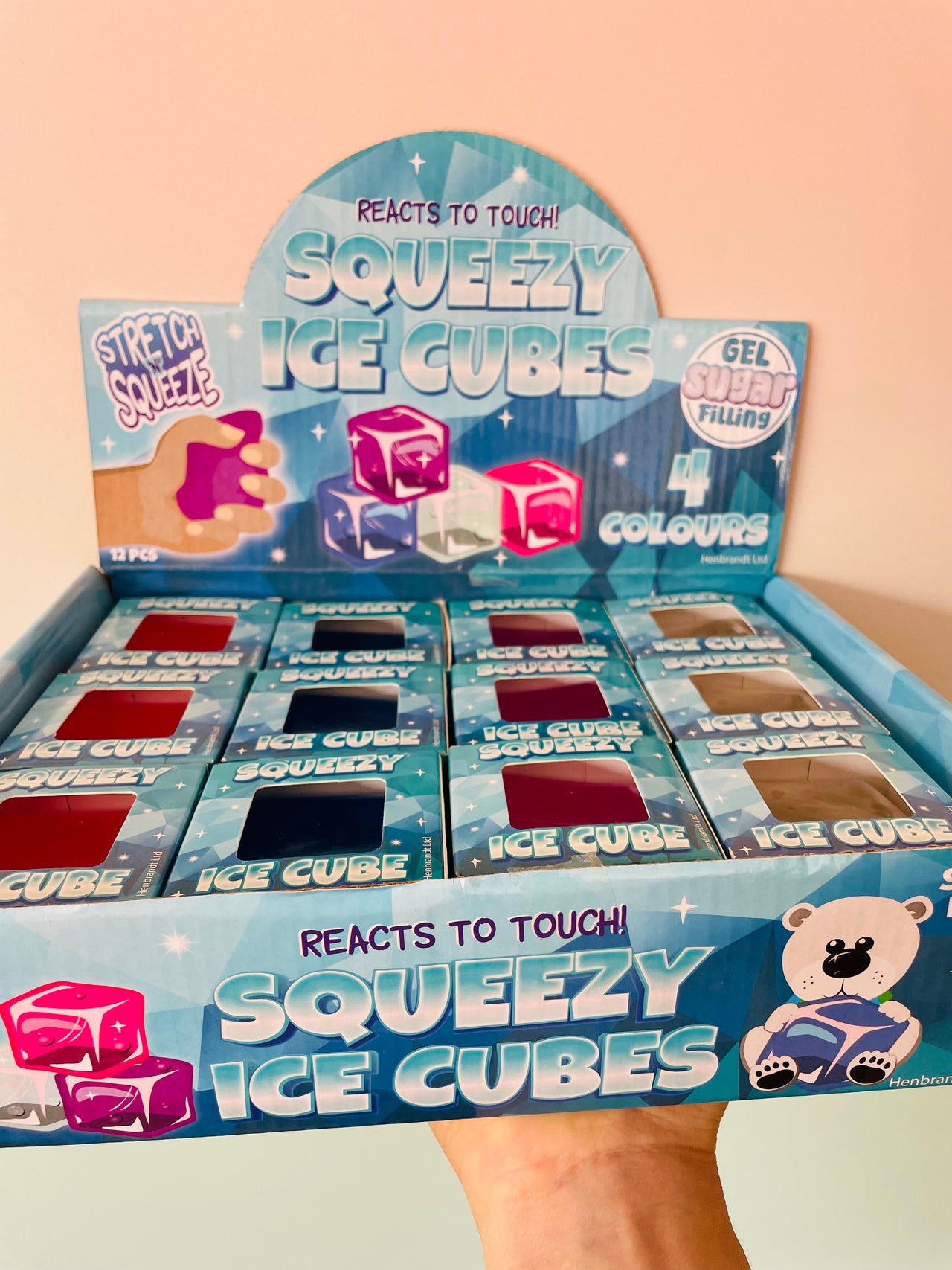 A hand holding a blue cardboard display box filled with small Squeezy Ice Cube packs. The packaging features the text 'REACTS TO TOUCH! SQUEEZY ICE CUBES' and shows gel ice cubes in different colors, including pink, blue, and red.