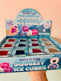 A hand holding a blue cardboard display box filled with small Squeezy Ice Cube packs. The packaging features the text 'REACTS TO TOUCH! SQUEEZY ICE CUBES' and shows gel ice cubes in different colors, including pink, blue, and red.