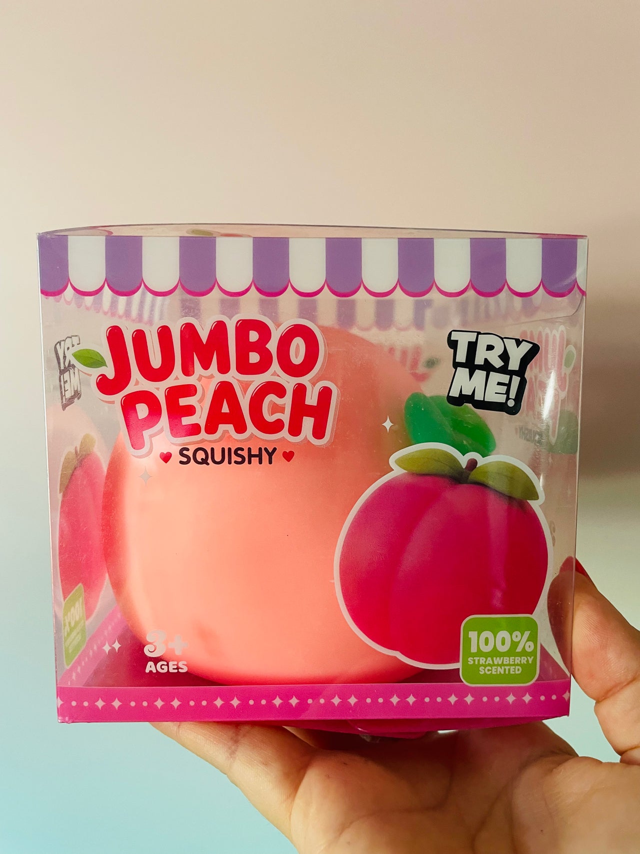 A hand holding a packaged squishy toy shaped like a peach, with the box reading 'JUMBO PEACH SQUISHY' and 'TRY ME!'.