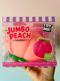 A hand holding a packaged squishy toy shaped like a peach, labeled 'JUMBO PEACH SQUISHY' with 'TRY ME!' and '100% STRAWBERRY SCENTED' text on the clear front packaging.