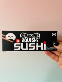 A hand with dark green nail polish holds a black box of Scrunchems Squishy Sushi against a pastel pink and light blue gradient background.