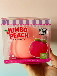 A hand holding a packaged squishy toy shaped like a peach, labeled 'JUMBO PEACH SQUISHY' with 'TRY ME!' and '100% STRAWBERRY SCENTED' on a clear plastic box.