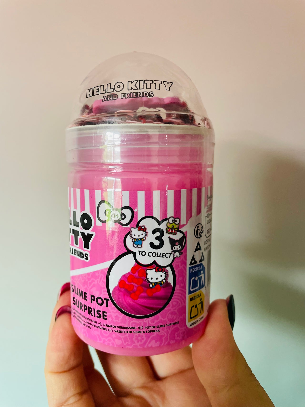 A hand holding a Hello Kitty Friends 3-in-1 slime pot surprise toy in a clear plastic tub with a pink label and a clear dome lid.