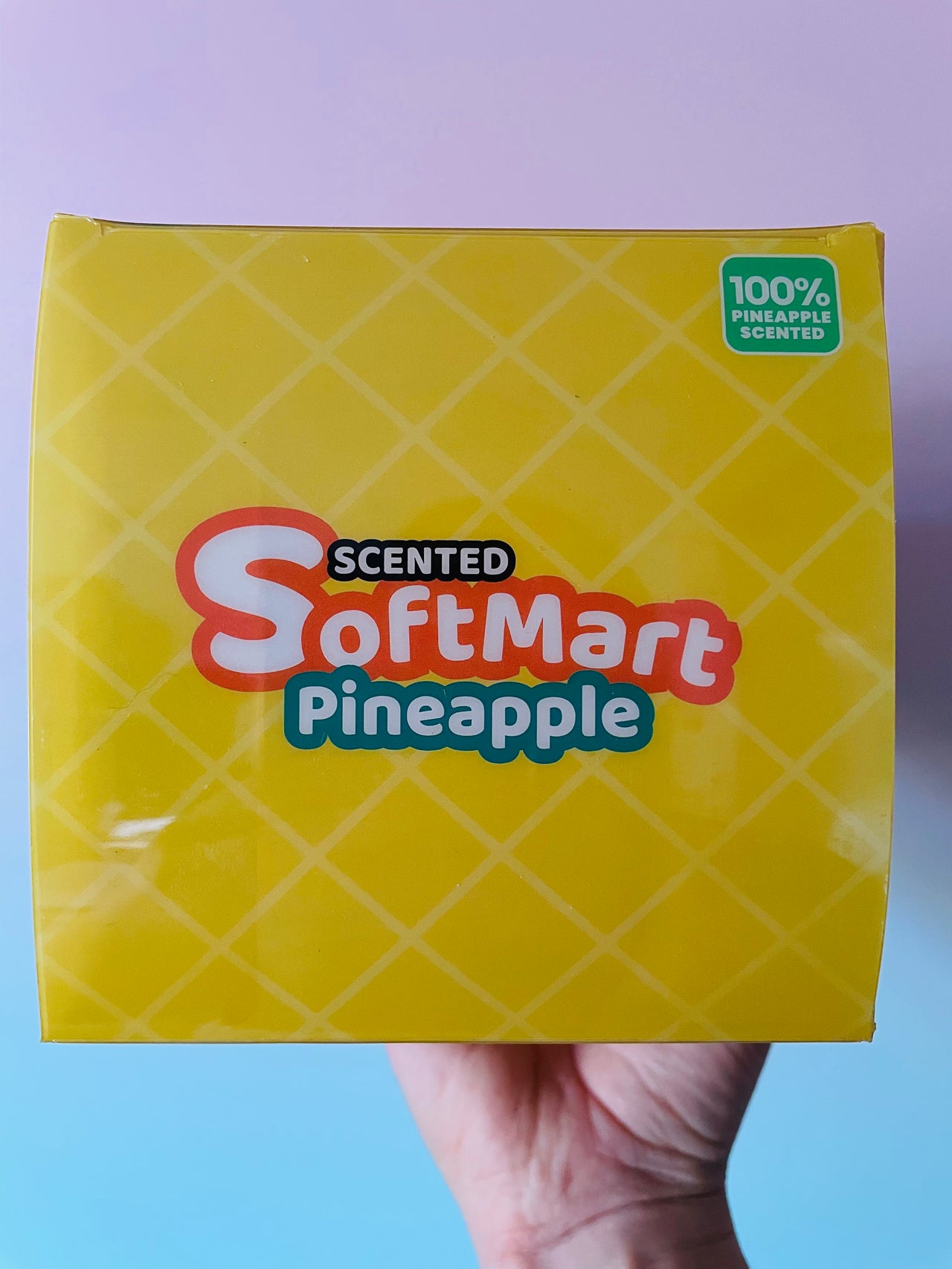 A hand holding a yellow box of Scented SoftMart Pineapple scented candles against a pastel purple and blue gradient background.