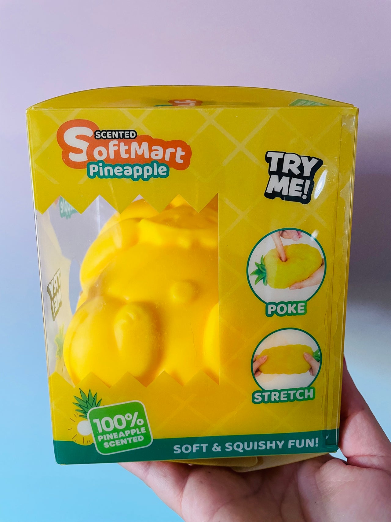 A hand holding a yellow box of SoftMart Scented Pineapple squishy toy against a pastel purple and blue background. The packaging features a clear window showing the yellow pineapple-shaped squishy inside, with text indicating 'TRY ME!', 'POKE', and 'STRETCH' features.