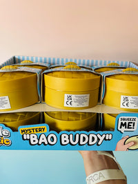 A retail display box filled with multiple yellow round squeeze toy containers labeled 'Mystery Bao Buddy,' with a hand holding the front display card against a pale pink and blue background.