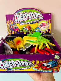 A display box filled with multiple packages of Creepsterz Stretchy Spider toys, held by a hand against a plain beige wall. The box contains several colorful, stretchy spider toys in yellow, green, red, and blue.