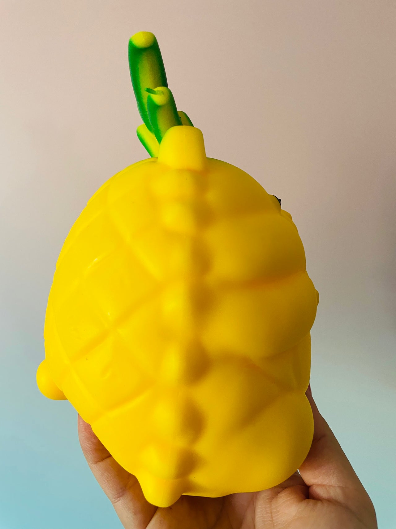 A hand holding a bright yellow, textured squishy toy shaped like a stylized fruit with a green stem, photographed against a soft beige-to-blue gradient background.