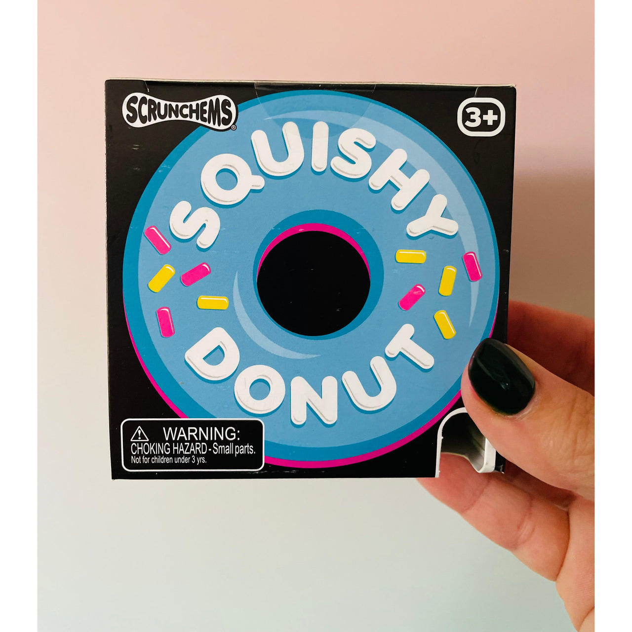 A hand with black nail polish holds a box of Scrunchems 'Squishy Donut' toys against a soft pink background. The packaging features a blue donut graphic with white lettering and pink and yellow sprinkles.