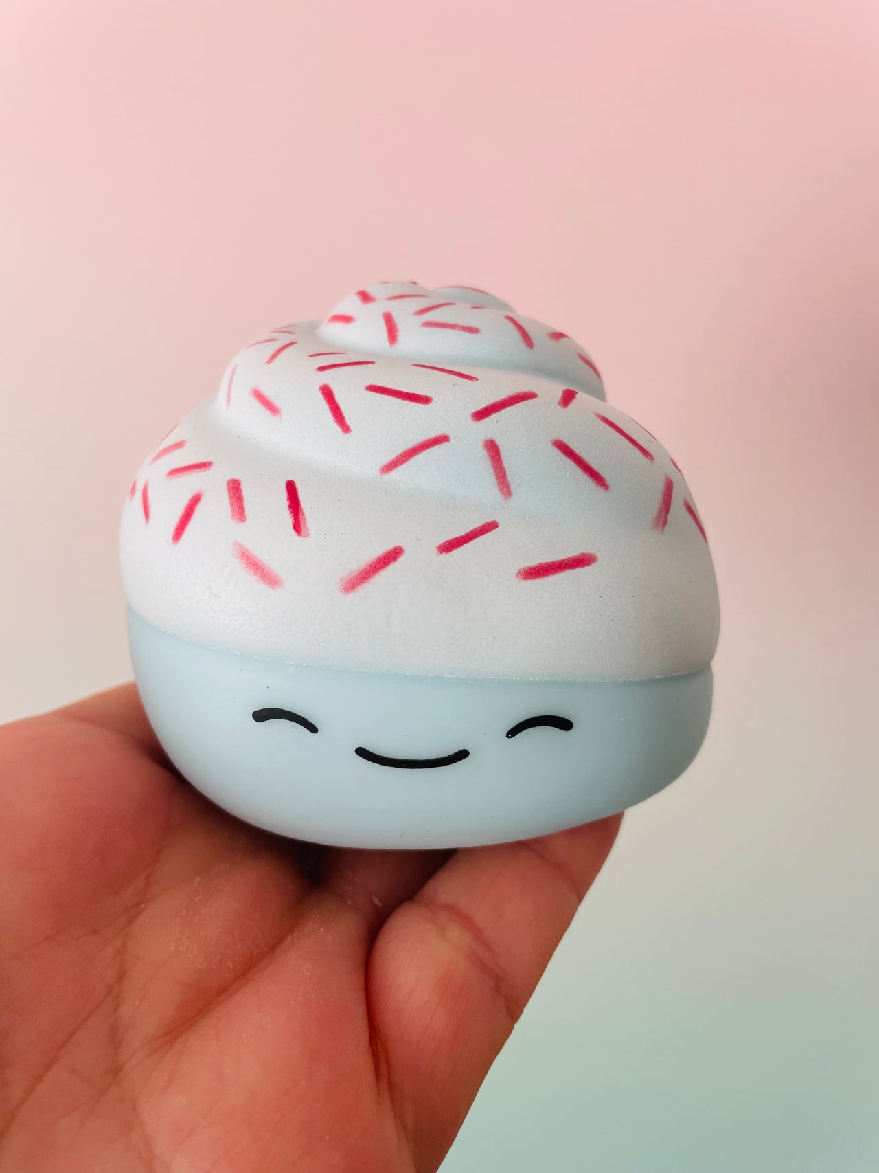 A hand holding a small, round, cupcake-shaped squishy toy with white frosting, red sprinkles, and a light blue base with a simple black smiling face.