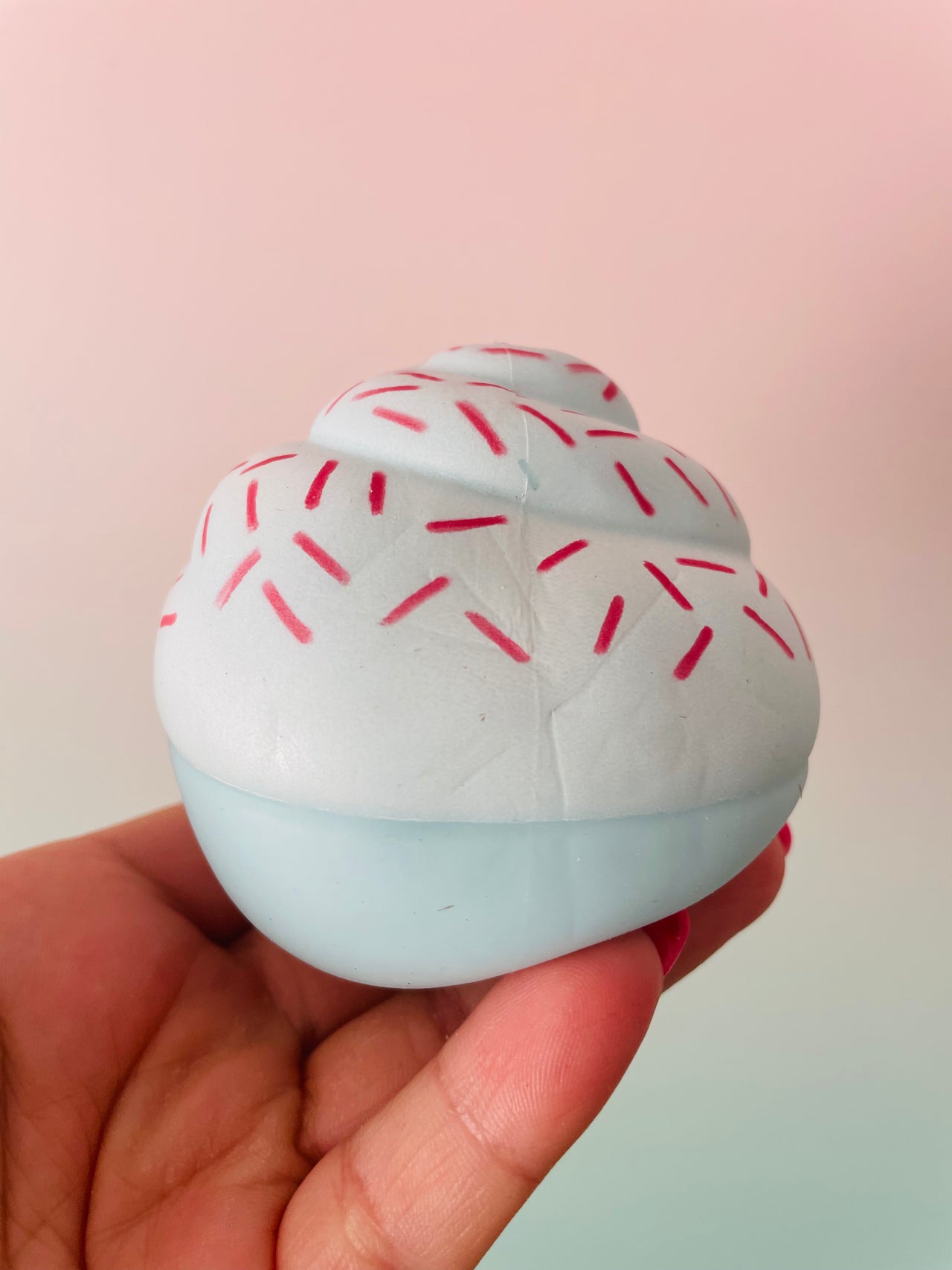A hand holding a small cupcake-shaped squishy toy with white frosting, red sprinkles, and a pale blue base against a soft pink and mint gradient background.