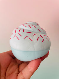 A hand holding a small cupcake-shaped squishy toy with white frosting, red sprinkles, and a pale blue base against a soft pink and mint gradient background.