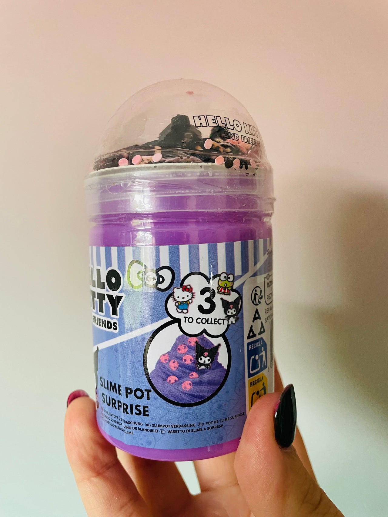 A hand holding a Hello Kitty Friends 'Slime Pot Surprise' collectible slime container. The purple plastic tub is filled with purple slime and decorated with pink and black glitter, topped with a clear dome lid.
