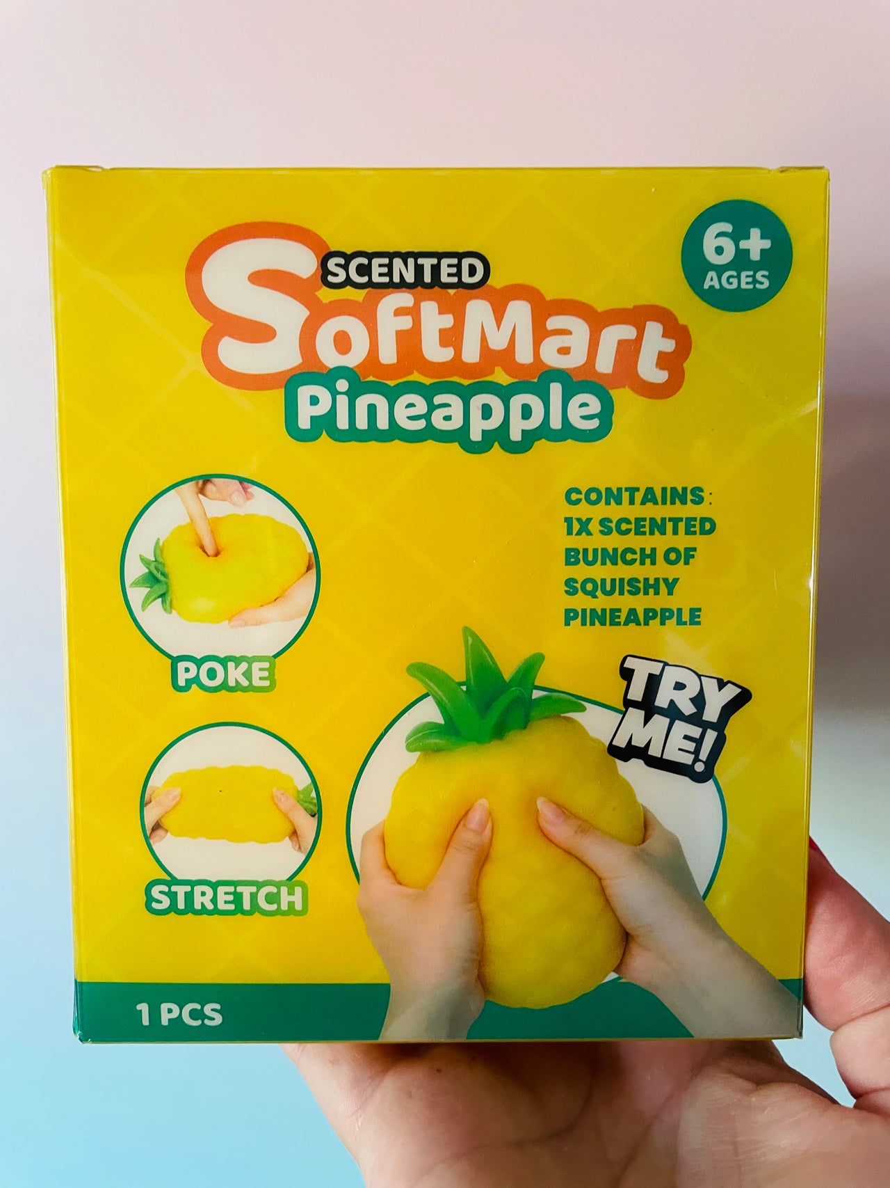 A hand holding a yellow box of Scented SoftMart Pineapple squishy toy. The packaging features a bright yellow background with green and orange branding, instructions on how to poke and stretch the pineapple, and a graphic of hands squeezing the yellow pineapple toy.