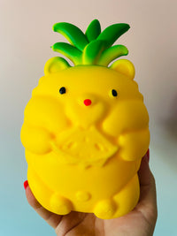 A yellow, bear-shaped pineapple squishy toy being held in a hand against a soft, light-colored background.