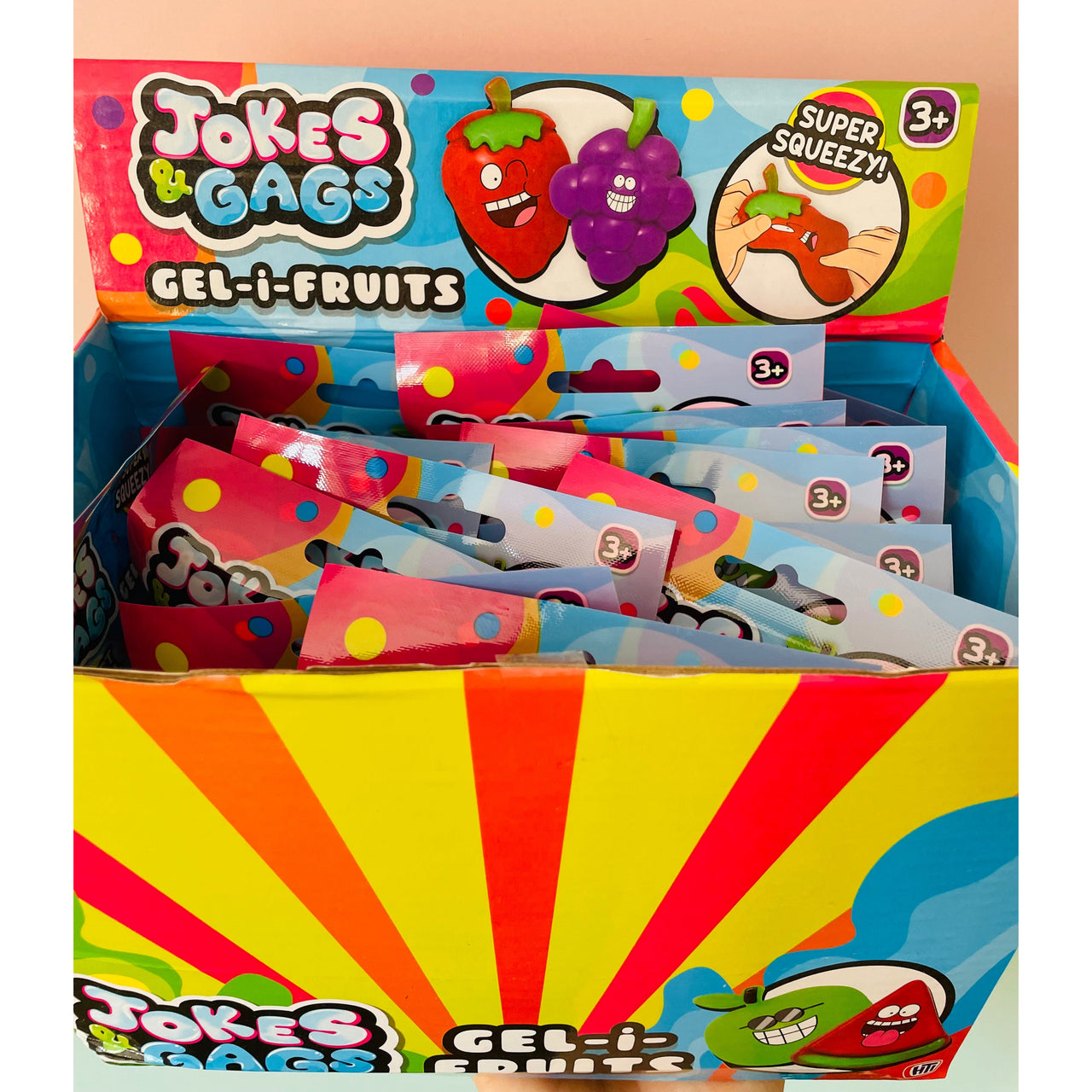 A retail display box filled with Jokes & Gags Gel-i-Fruits blind bag packs. The packaging features bright, colorful graphics with cartoon fruit characters and a yellow and red sunburst pattern on the front.