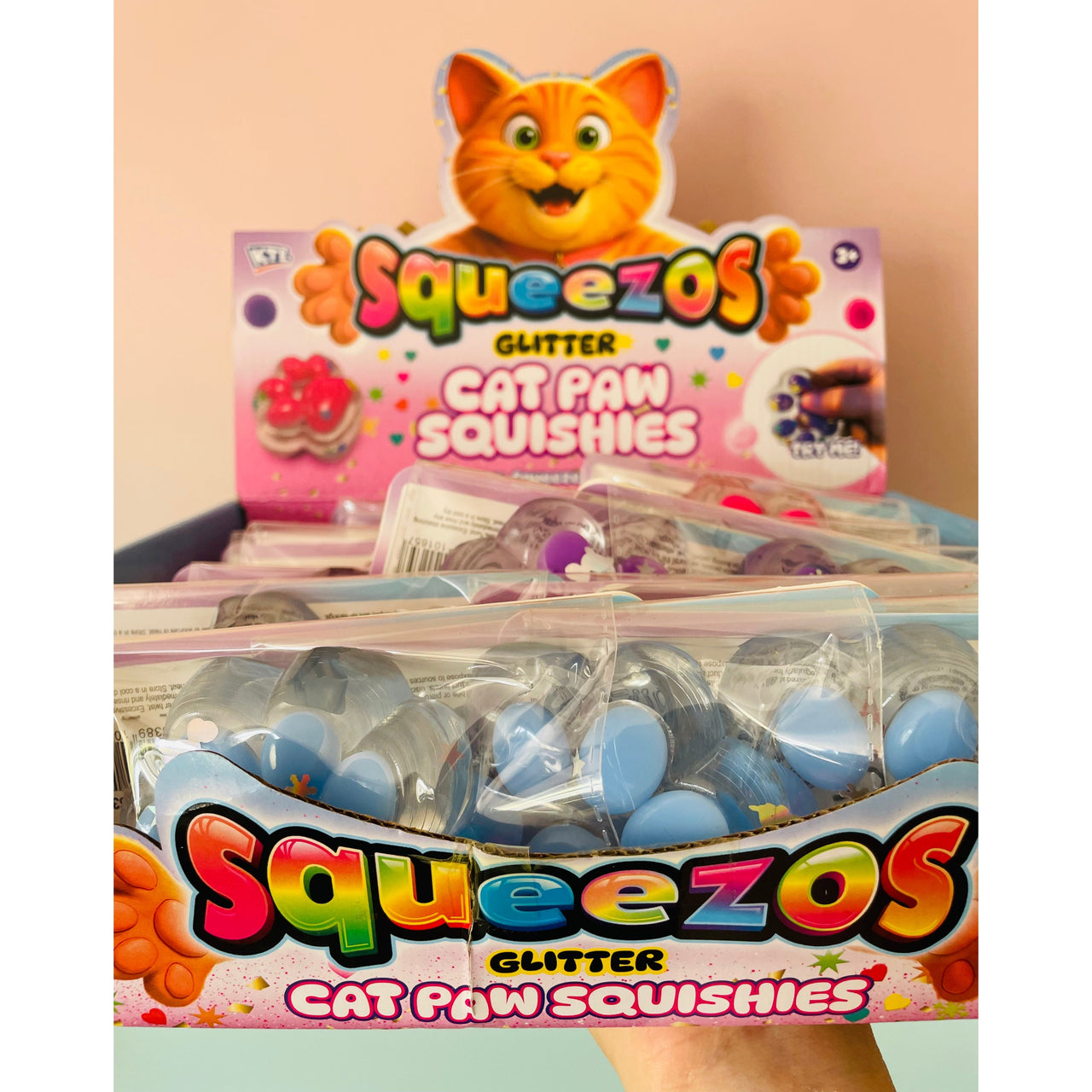 A display box of Squeezos Glitter Cat Paw Squishies, showing multiple small blue glitter squishies inside clear plastic bags. The packaging features a cartoon orange cat face and colorful 'Squeezos' branding, set against a plain beige background.