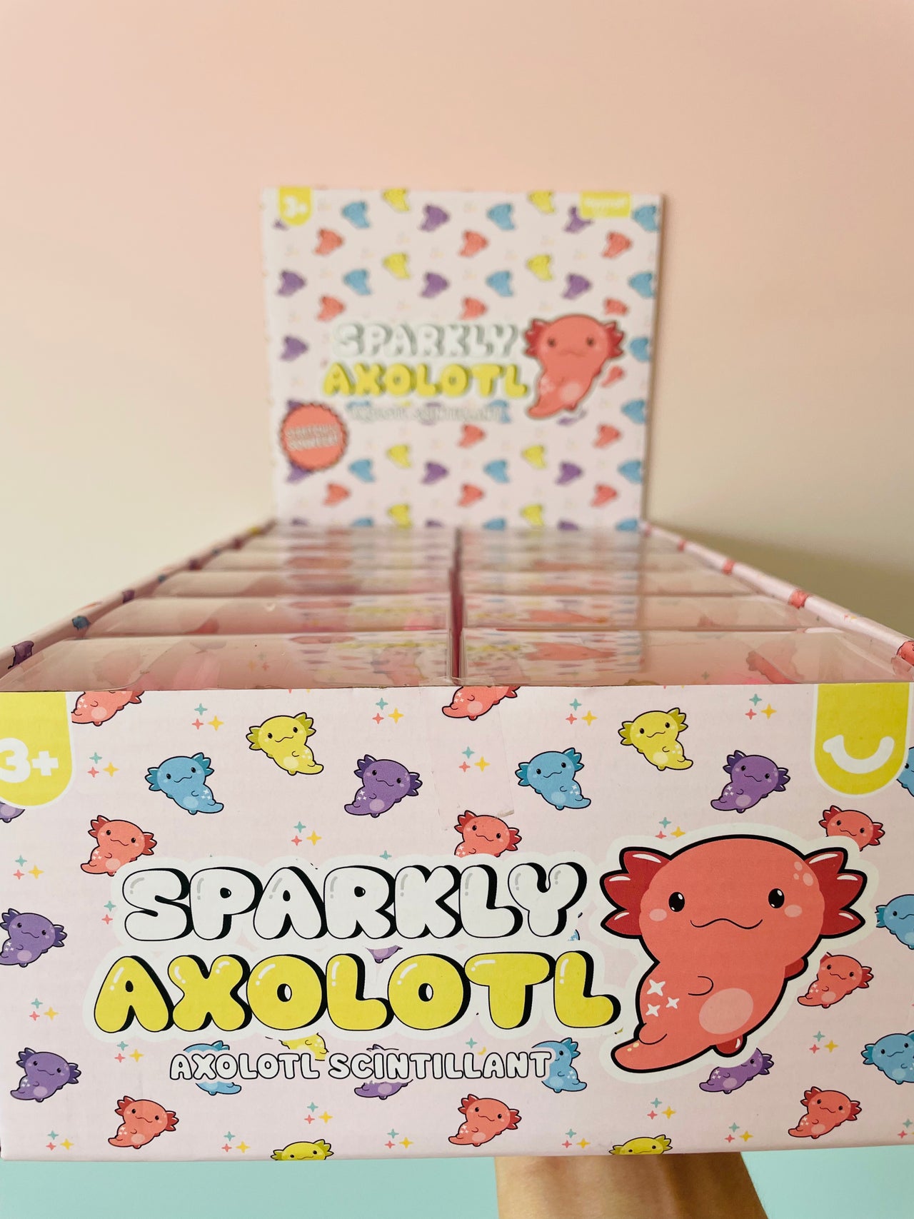 A retail display box filled with small packaged items labeled 'SPARKLY AXOLOTL' and a matching upright card or package insert, shown against a soft peach background.