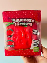 A hand holding a packaged novelty toy shaped like a strawberry, labeled 'Squeeze Strawberry,' with a red body, clear plastic window, and printed instructions on the front.