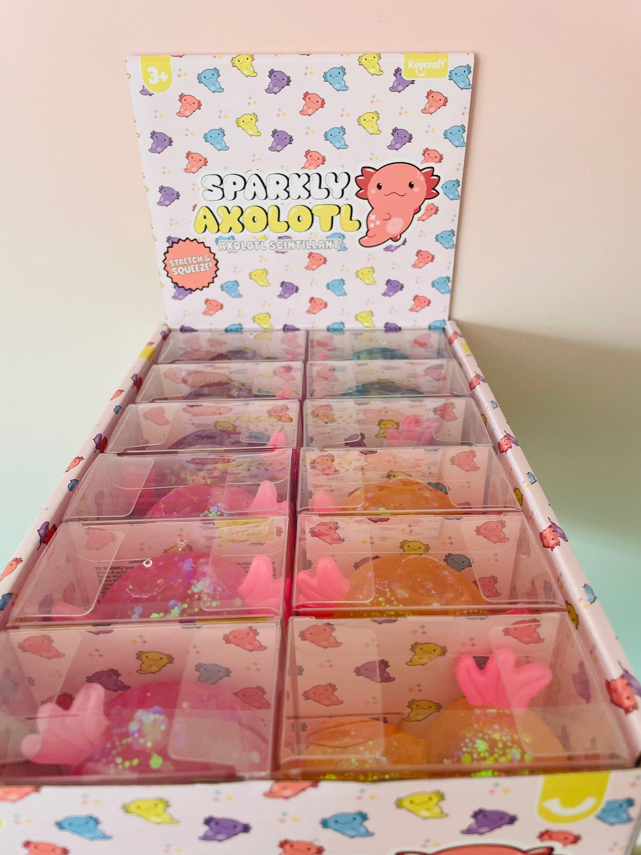 An open display box of 'Sparkly Axolotl' slime charms, showing multiple clear plastic compartments filled with pink, orange, and purple sparkly slimes, with a branded cardboard backing.