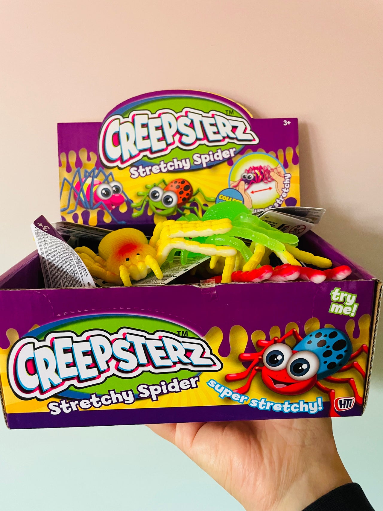 A hand holding a purple Creepsterz Stretchy Spider candy box filled with colorful stretchy toy spiders, against a plain light beige wall.