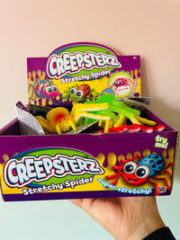 A hand holding a purple Creepsterz Stretchy Spider candy box filled with colorful stretchy toy spiders, against a plain light beige wall.