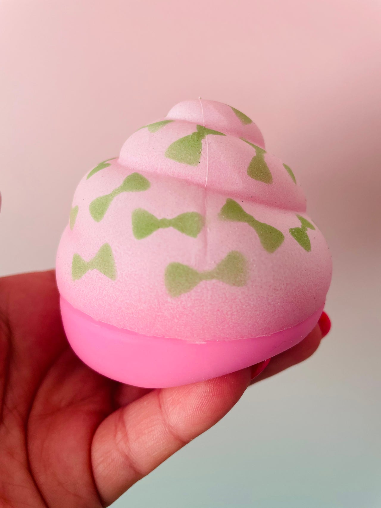 A hand holding a pink cupcake-shaped squishy toy with green bow tie patterns on a soft pink background.