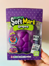 A hand holding a purple box of Scented Soft Mart Grapes toy. The packaging features a clear window showing a purple, squishy grape-shaped toy inside.