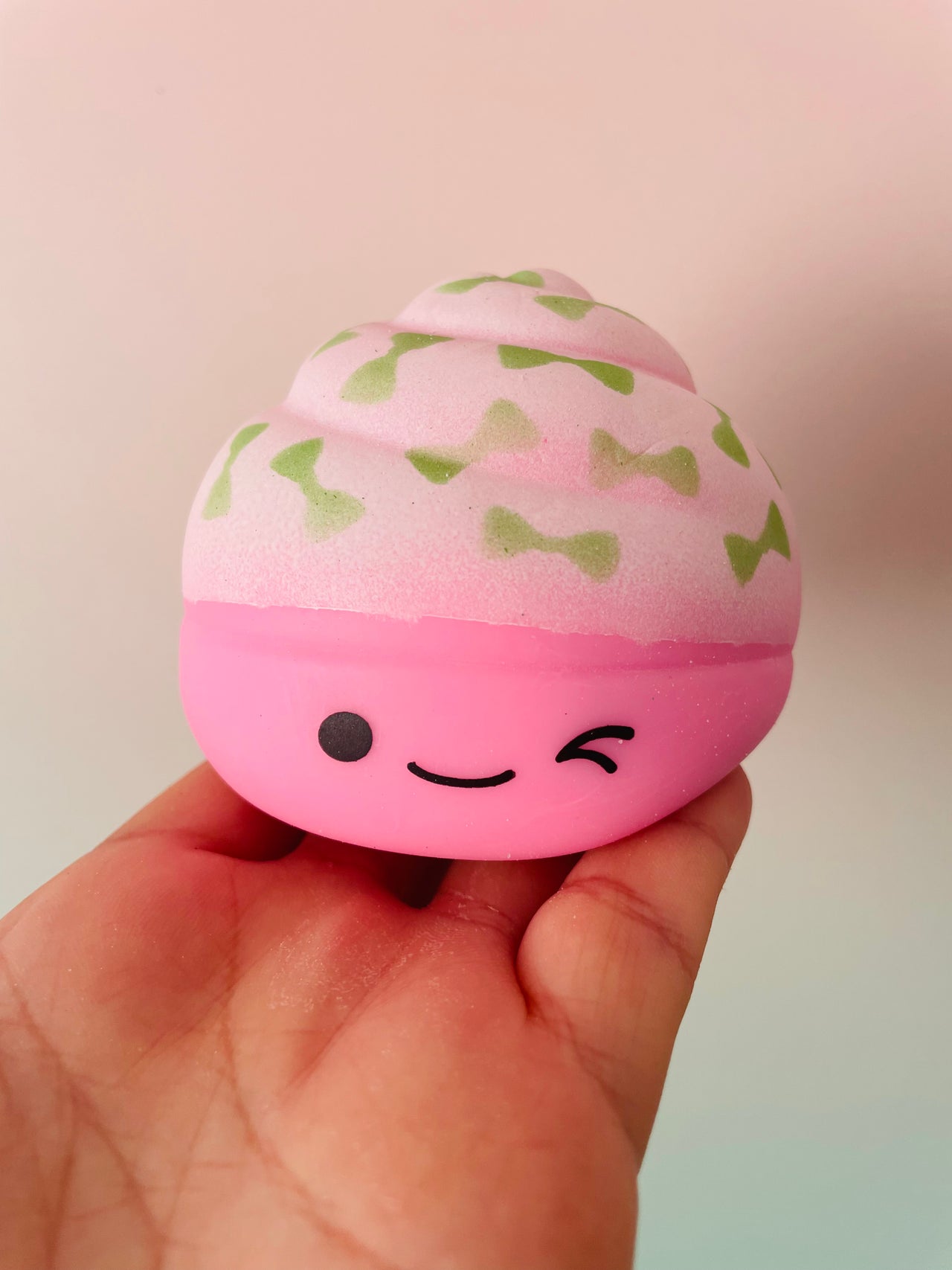 A hand holding a pink squishy toy shaped like a cupcake, with a winking face and green bow-tie patterns on a pink frosting-like top, against a soft pastel background.