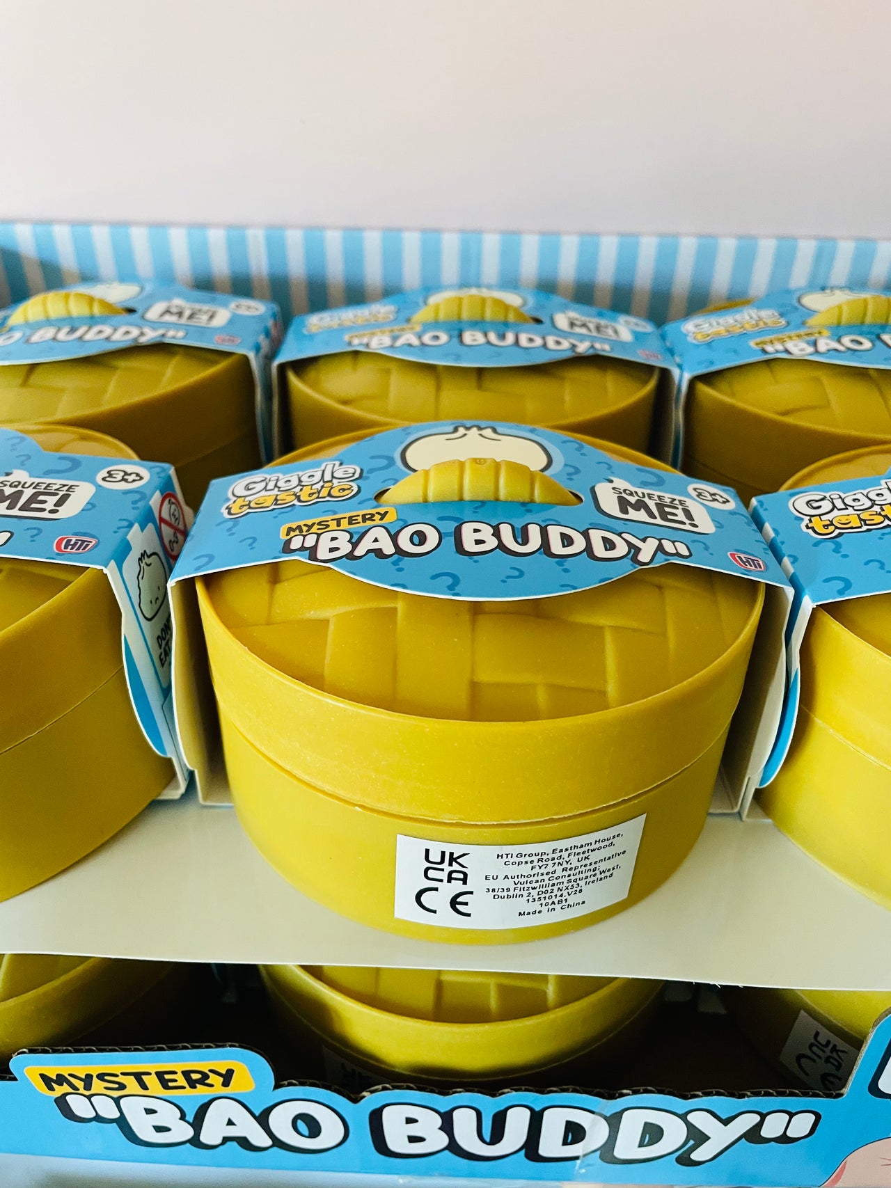 A display of yellow mystery toy containers labeled 'Mystery Bao Buddy' on retail shelves. The containers are packaged with blue and white labels, and multiple units are visible on the shelves.