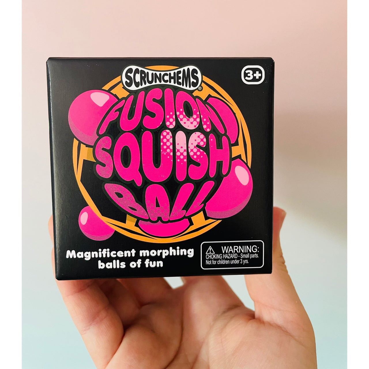 A hand holding a black box of Scrunchems Fusion Squish Ball, featuring bright pink and orange lettering, against a soft pastel pink and blue background.